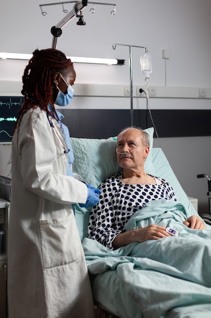 Praticien médical portant un masque chirurgical dans la chambre d'hôpital lors de la consultation d'un homme âgé malade et malade, respirant à l'aide d'un tube à essai et d'une perfusion intraveineuse attachée