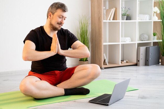 Pratique du yoga Homme méditant Formation en ligne Vidéoconférence Un homme souriant et paisible en tenue de sport décontractée assis pose de lotus tenant les mains de namaste à la recherche d'un ordinateur portable à l'intérieur de la maison