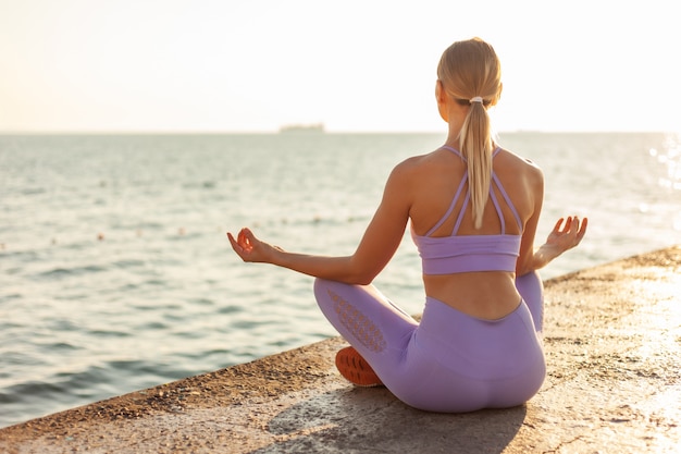 Pratiquer le yoga. Méditation au lever du soleil. Jeune femme mince assise en position du lotus sur la plage. Concept de mode de vie sain