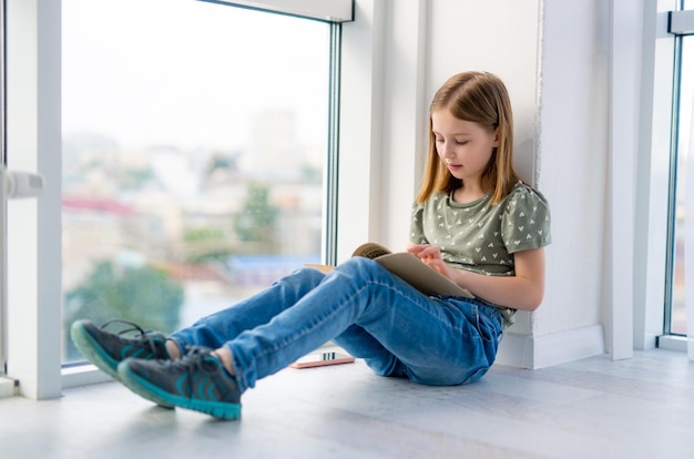 Préadolescente assise sur le rebord de la fenêtre et lisant un livre en papier avec la lumière du jour belle écolière lea...