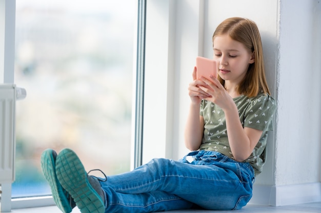 Préadolescente assise sur le rebord de la fenêtre avec un smartphone et envoyant des SMS à une belle écolière féminine en train de se préparer...