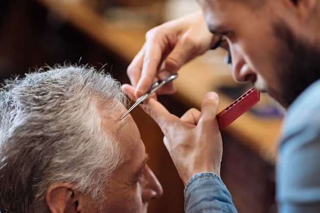 Précis et propre. Gros plan d'un homme senior sympa se faire couper les cheveux par un coiffeur professionnel alors qu'il était assis au salon de coiffure.