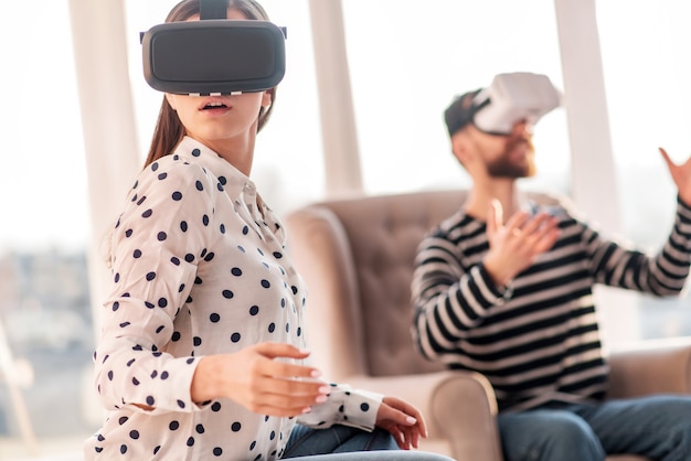 Premier coup d'oeil. Belle femme élégante sincère assise sur l'arrière-plan flou tout en se tournant vers la caméra et en expérimentant avec un casque VR