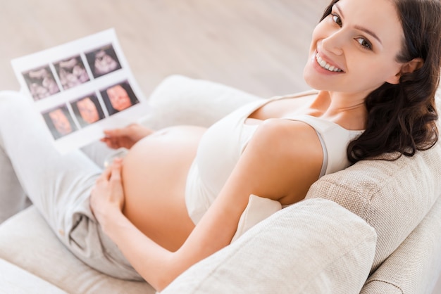 Première photo de mon bébé. Vue de dessus d'une femme enceinte heureuse assise sur un canapé et tenant une image radiographique de son bébé