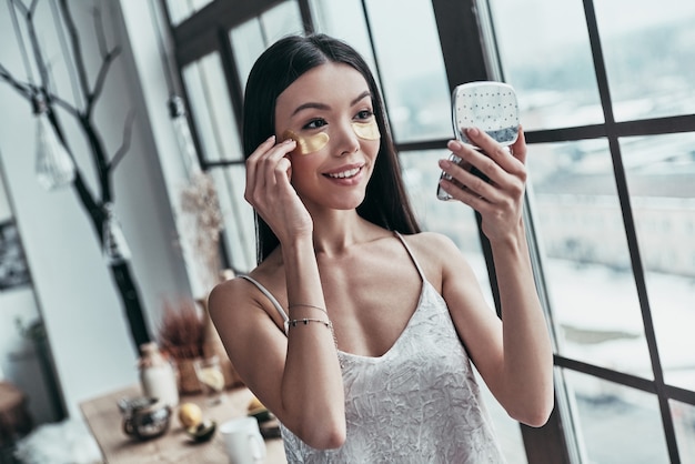 En prenant bien soin. Jolie jeune femme utilisant des cache-œil et souriant tout en passant du temps à la maison