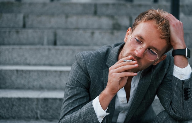 Prend une pause et fume une cigarette Jeune homme d'affaires en tenue de soirée grise est à l'extérieur de la ville