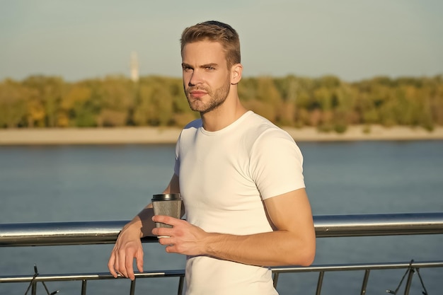 Prendre un café avec vous Jeune homme boit du café à l'extérieur Bel homme tient une tasse à emporter Homme célibataire sur la berge Homme mal rasé dans un style décontracté Garde-robe d'été Mode et style