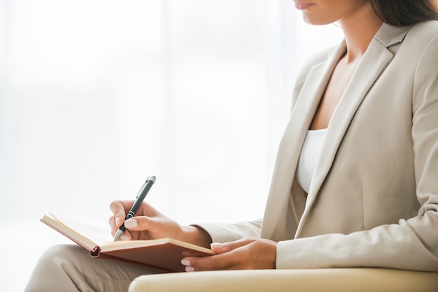 Prendre des notes d'affaires. Vue latérale d'une jeune femme d'affaires en costume écrivant quelque chose dans un bloc-notes alors qu'il était assis à la chaise