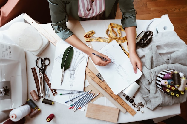 Prendre de nouvelles idées sur papier. Vue de dessus d'une fabuleuse jeune femme tailleur avec machine à coudre pense à une nouvelle robe. Les articles sur mesure sont à table. Ciseaux, ruban à mesurer, règle, cordes