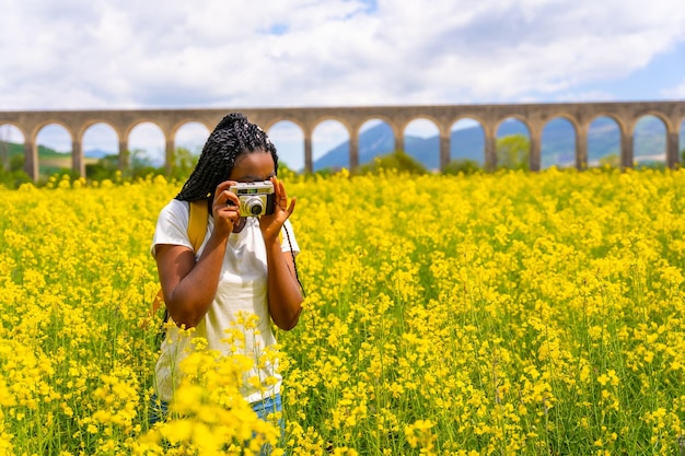 Prendre des photos avec un appareil photo vintage une fille ethnique noire avec des tresses un voyageur dans un champ de fleurs jaunes