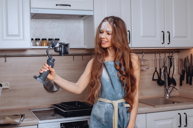 Prendre des photos de soi sur les réseaux sociaux. Une femme au foyer pose pour l'appareil photo en tenant son téléphone sur un trépied et en souriant vivement. Tablier de maison légèrement taché de farine. Séance photo à domicile dans la cuisine.