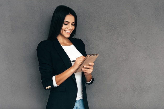 Prendre quelques notes. Jolie jeune femme en vêtements décontractés intelligents écrivant quelque chose dans son cahier et souriant en se tenant debout sur fond gris