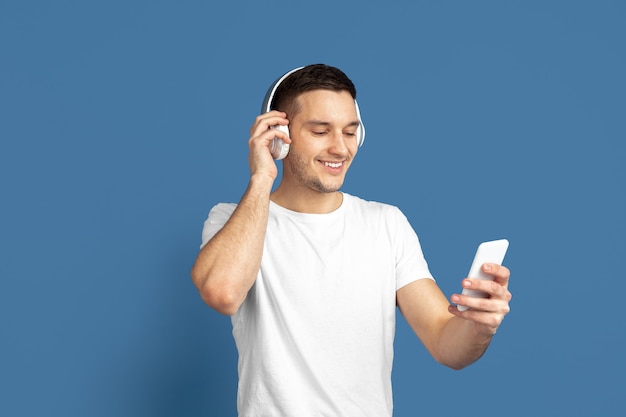 Prendre selfie, écouter de la musique. Portrait de jeune homme caucasien sur fond bleu studio. Beau modèle masculin dans un style décontracté, couleurs pastel. Concept d'émotions humaines, d'expression faciale, de ventes, d'annonces.