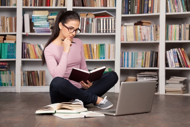 Préparation aux examens. Jeune femme réfléchie tenant un livre et regardant l'ordinateur portable alors qu'elle était assise par terre à la bibliothèque