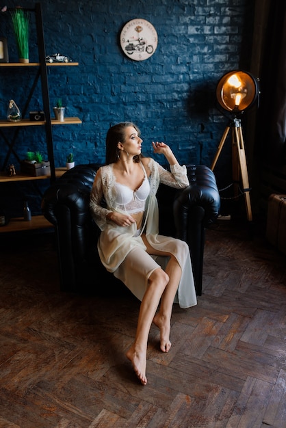 Préparation du matin de la mariée. Belle jeune femme en déshabillé de mariage blanc dans un studio sombre