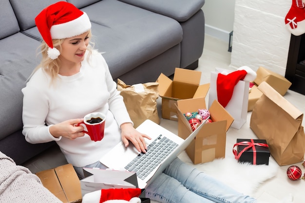 Préparation de la fête de Noël. Femme commandant des cadeaux et des décorations sur un ordinateur portable, assise parmi des coffrets cadeaux et des colis, espace de copie