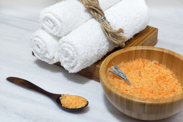 Préparation pour la procédure de spa - sel de bain orange dans un bol en bois et cuillère et trois serviettes blanches sur une boîte en bois sur une table en marbre blanc