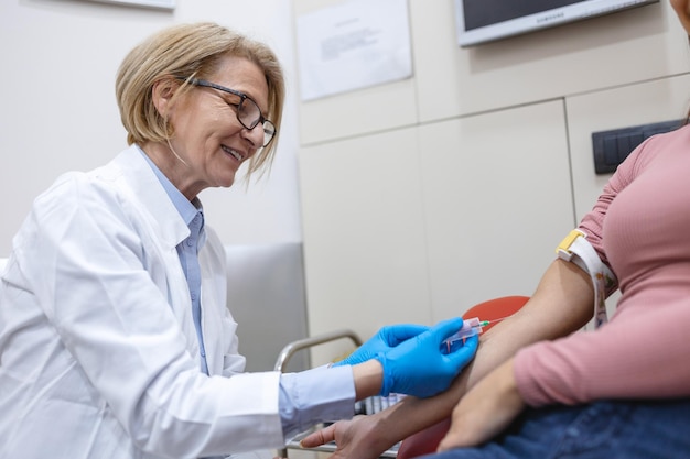 Préparation pour le test sanguin avec une jolie jeune femme par une femme médecin uniforme médical sur la table dans une pièce blanche et lumineuse L'infirmière perce la veine du bras du patient avec un tube vierge d'aiguille