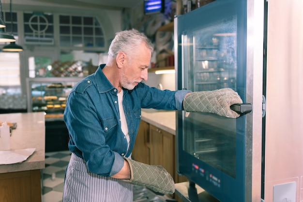 Près du four. Propriétaire de boulangerie aux cheveux gris portant une chemise en jean debout près du four à la recherche de baguettes