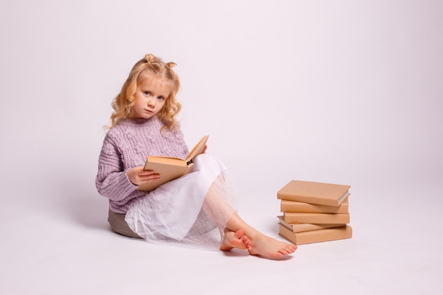 Preschool girl blonde assise sur un fond blanc avec un livre dans ses mains, souriant, pensant