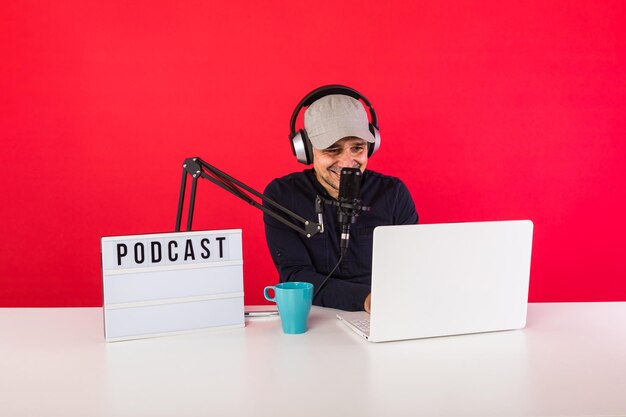 Présentateur masculin avec casquette dans un studio d'enregistrement radio podcast souriant et consultant Internet, à côté d'un ordinateur, d'un microphone et d'un caisson lumineux avec le mot podcast, sur fond rouge.