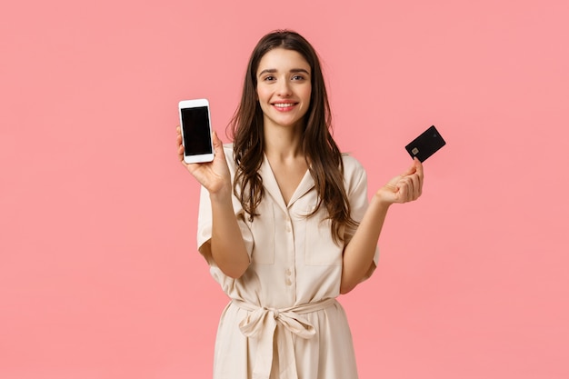 Présente, concept de shopping et de beauté. À la mode jeune femme en robe à la mode, tenant un smartphone et une carte de crédit, souriant joyeusement, commandez un produit en ligne, debout rose