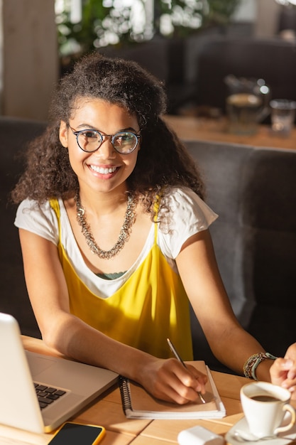 Prêt à écrire. Une femme positive et ravie qui vous sourit en étant assise avec son stylo à la main