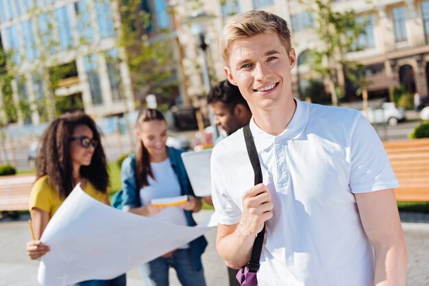 Prêt à partir. Blonde positive gardant le sourire sur son visage et tenant le sac à dos tout en posant devant la caméra