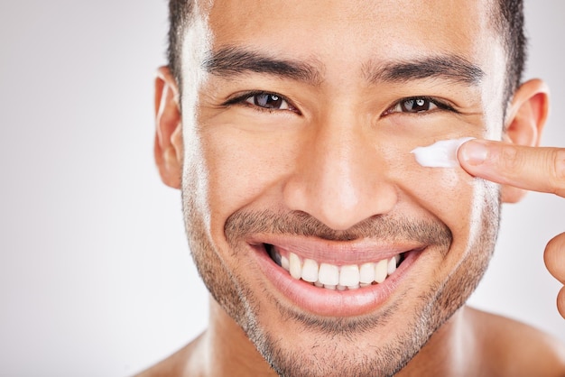 Prêt pour l'appareil photo Gros plan portrait en studio d'un jeune homme appliquant joyeusement une crème hydratante sur son visage sur un fond gris