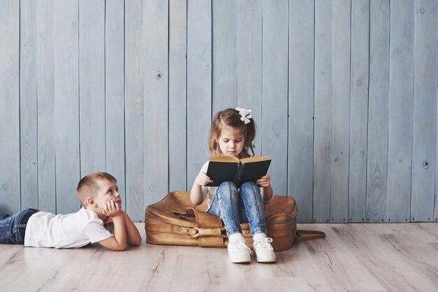 Prêt pour les grands voyages. Bonne petite fille et garçon lisant un livre intéressant portant une grosse mallette et souriant. Voyage, liberté et imagination