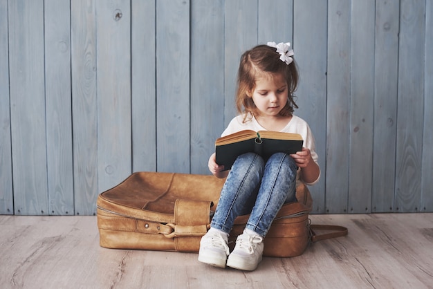 Prêt pour les grands voyages. Bonne petite fille lisant un livre intéressant portant une grosse mallette et souriant. Voyage, liberté et imagination