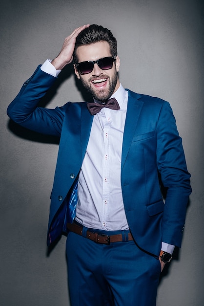 Prêt pour un rendez-vous. Beau jeune homme gai en costume gardant la main dans les cheveux et regardant la caméra avec le sourire en se tenant debout sur fond gris