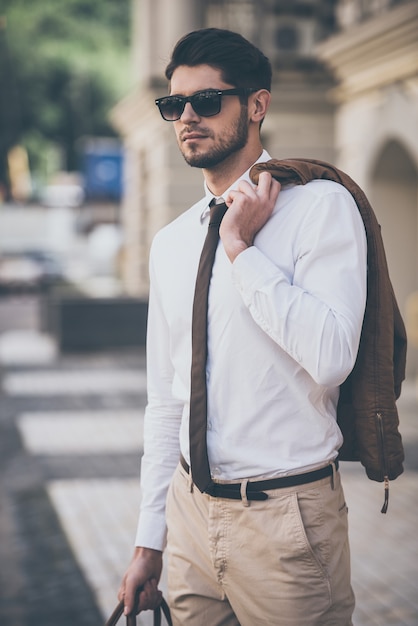 Prêt pour un rendez-vous. Beau jeune homme à lunettes de soleil tenant une veste et détournant les yeux en marchant à l'extérieur