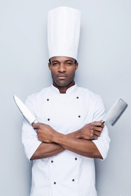 prêt à travailler. jeune chef africain confiant en uniforme blanc tenant des couteaux dans ses mains