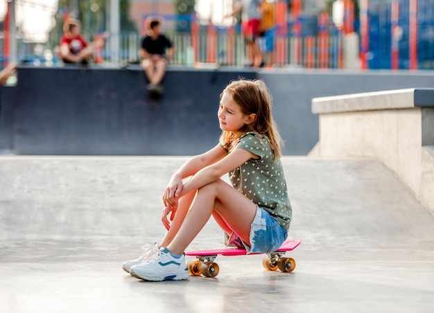 Preteen girl sitting on skateboard à l'extérieur et regardant en arrière. Enfant de patineuse près de la rampe d'équitation de la ville