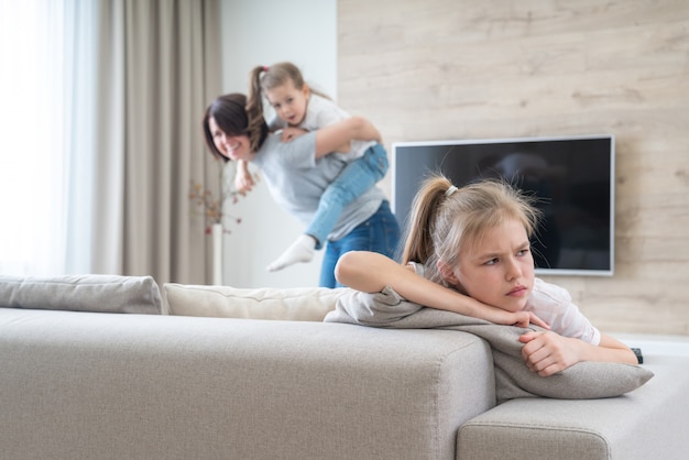 Preteenage triste fille assise sur un canapé pendant que la mère s'amuse avec la sœur, le concept de la jalousie