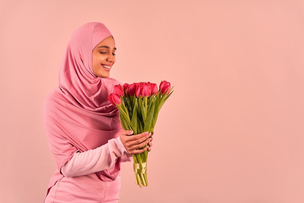 Printemps et Journée internationale de la femme Une belle jeune femme musulmane vêtue d'un hijab et d'une robe rose tendre tient un bouquet de tulipes et les regarde