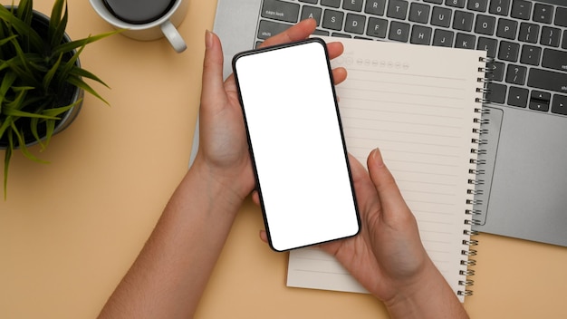 Prise de vue aérienne Une main féminine tenant une maquette d'écran blanc de smartphone sur un bureau minimal