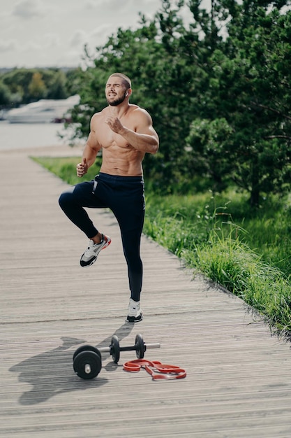 Prise de vue en extérieur d'un bel homme barbu musclé en forme fait un exercice de tapotement du genou élevé se réchauffe avant de courir a un entraînement cardio utilise des équipements de sport a un corps fort fait preuve de détermination