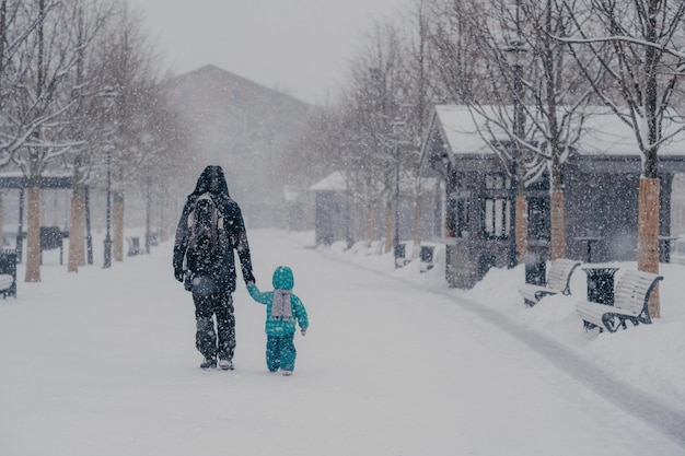 Prise de vue en extérieur d'un petit enfant et d'un père couvrant la distance étant sur le chemin de la maison se tenir la main profiter du temps neigeux d'hiver Vue arrière des personnes marchant dans la rue pendant les chutes de neige Concept de saison froide