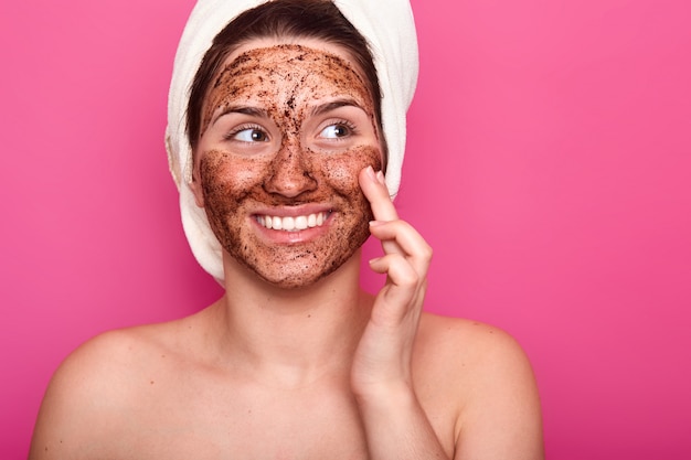 Prise de vue en intérieur d'une jeune femme applique un masque cosmétique pour le visage, a un sourire à pleines dents, a une serviette enveloppée sur la tête, pose à moitié nue