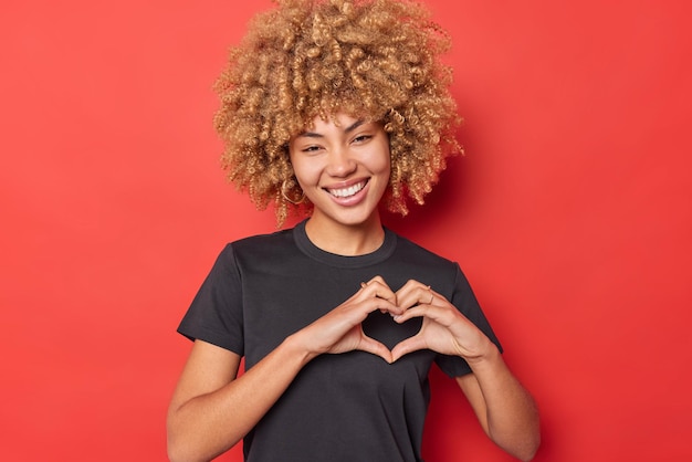 Prise de vue en intérieur d'une jeune femme romantique assez souriante forme le cœur avoue amoureux porte un t-shirt noir décontracté isolé sur fond rouge Concept de relation Je t'aime signe Be my valentine