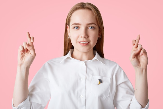 Prise de vue en intérieur d'une jolie femme aux yeux bleus aux cheveux longs, garde les doigts croisés, souhaite que les rêves deviennent réalité, isolé sur le mur rose Concept de langage corporel