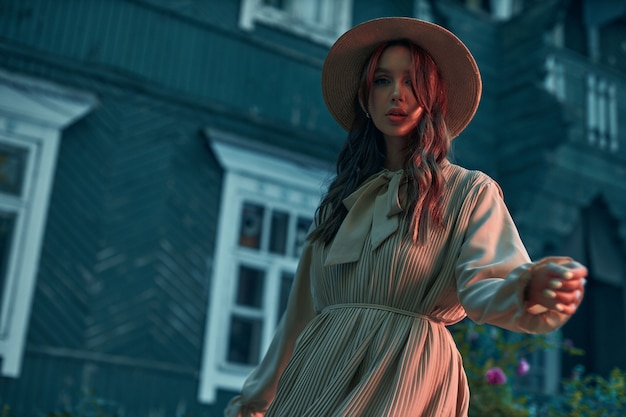 Prise de vue moderne dans des tons bleus, une belle jeune fille vêtue d'une robe d'été et d'un chapeau de paille se tient près d'une vieille maison et regarde mystérieusement la photo de haute qualité de l'appareil photo