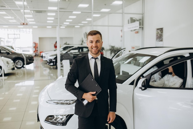 Une prise de vue rapprochée d'un assistant de magasin caucasien heureux, confiant et souriant tenant un bloc-notes dans des vêtements formels et regardant la caméra dans un magasin de concessionnaires d'automobiles.