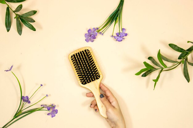 prise de vue rapprochée de la main de la femme tenant une brosse à cheveux en bois avec des fleurs violettes violettes et une feuille de plante sur le dos rose