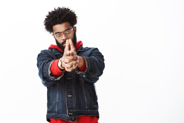 Prise de vue en studio d'un afro-américain séduisant et élégant dans des verres tirant les mains devant avec le geste du pistolet comme s'il tenait un pistolet en ayant l'air audacieux de viser la cible sur un mur gris