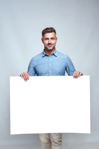 Prise de vue en studio d'un bel homme barbu dans des vêtements décontractés tenant un tableau blanc vide et souriant en se tenant debout sur fond gris. Publicité