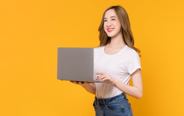 Prise de vue en studio d'une belle femme asiatique joyeuse en t-shirt blanc et tenant un ordinateur portable en tapant sur le clavier sur fond jaune.