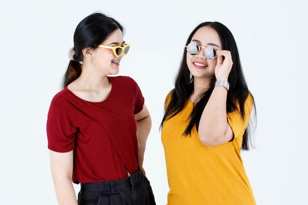 Prise de vue en studio de deux jeunes amies asiatiques à la mode et élégantes en tenue de t-shirt décontractée, portant des lunettes de soleil vintage à la mode, debout, souriantes, se regardant les yeux sur fond blanc.